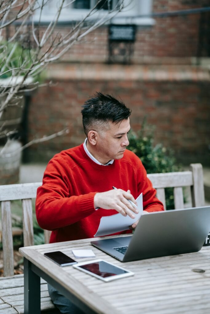 A man in a red sweater works on paperwork and a laptop at a wooden table outdoors.